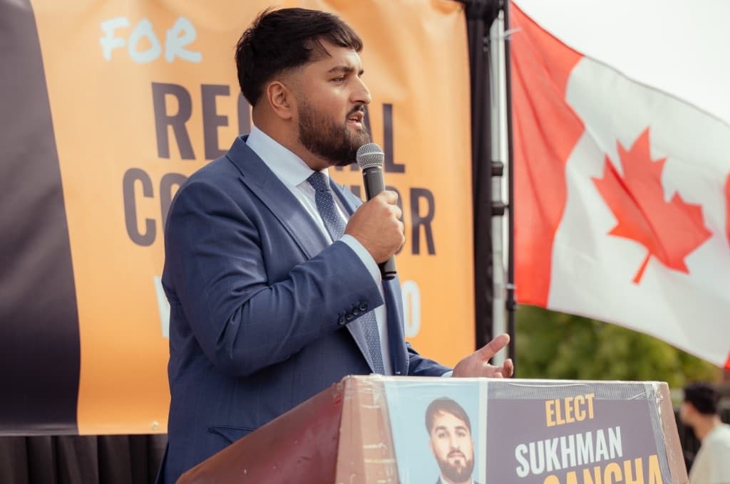 Sukhman Sangha speaking at a campaign podium beside a Canadian flag